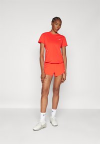 Piros sportos póló és rövidnadrág, könnyű anyagból készült. Nike logó díszíti. Fehér zoknikkal és világos sportcipőkkel van párosítva.