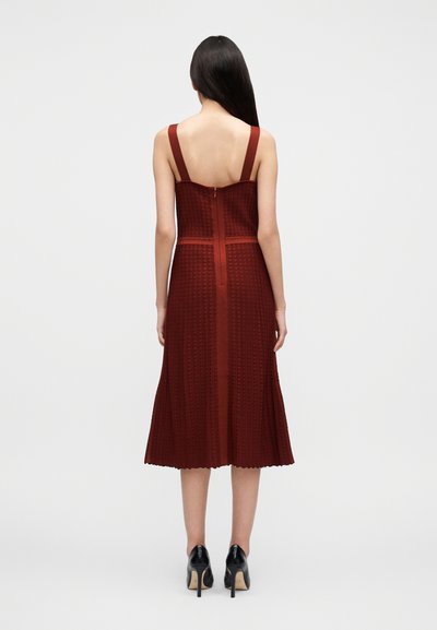 Μπορντό φόρεμα με υφή σχεδίου, φαρδιές τιράντες και φερμουάρ στο πίσω μέρος. Καμπυλωτό τελείωμα με κυματιστές άκρες, σε συνδυασμό με μαύρα παπούτσια με τακούνι.