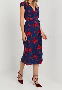 Kvinna iklädd en midiklänning i marinblått med röd blommig tryck och burgundy högklackade sandaletter, står mot en enkel vit bakgrund.