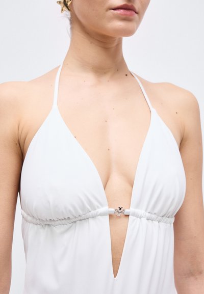 Γυναίκα που φοράει λευκό φόρεμα με λαιμόκοψη halter και βαθύ V ντεκολτέ, με μικρό ασημένιο κούμπωμα στο κέντρο μπροστά.