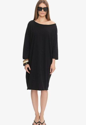 Kobieta w przesadnie dużej czarnej sukience z odkrytymi ramionami, dużych czarnych okularach przeciwsłonecznych, czarnych klapkach i nałożonych jedna na drugą bransoletkach na lewym nadgarstku, stojąca.