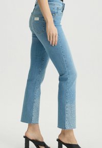Jeans in denim blu chiaro con taglio a zampa d'elefante, arricchiti da abbellimenti in strass lungo l'orlo e finitura grezza.