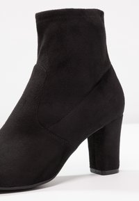 Bottine en daim noir avec un talon carré, une texture lisse, des panneaux latéraux incurvés et un détail de couture minimaliste.