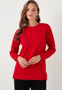 Maglietta rossa a maniche lunghe realizzata in tessuto morbido, con collo tondo, vestibilità rilassata, rifinita con polsini e orlo a costine, abbinata a pantaloni neri.