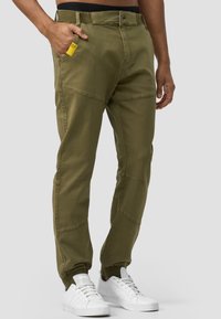Pantalones verde oliva con un ajuste entallado, hechos de tela ligera, que cuentan con bolsillos laterales y puños de canalé en los tobillos. Llevados con zapatillas blancas.