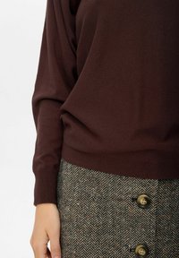 Brauner Strickpullover mit langen Ärmeln, kombiniert mit einem Rock in Fischgrätmuster, der große gelbe Knöpfe hat. Weicher Stoff, figurbetonter Bund.