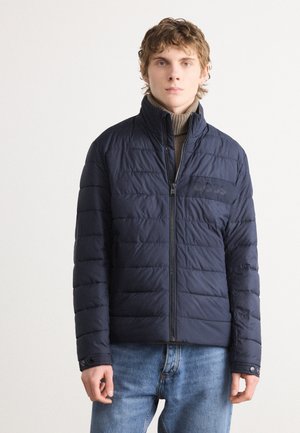 Temno modra puffer jakna z zadrgo spredaj, vertikalno prešita tekstura, visok ovratnik in detajl logotipa na prsih. Nosi se nad bež puloverjem z visokim ovratnikom.