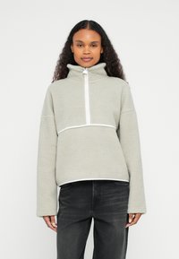 Jasnozielona bluza polarowa z wysokim kołnierzem, suwakiem do ćwiartki i białym akcentem pod klatką piersiową oraz przy dolnym brzegu. Miękka, teksturowana tkanina.