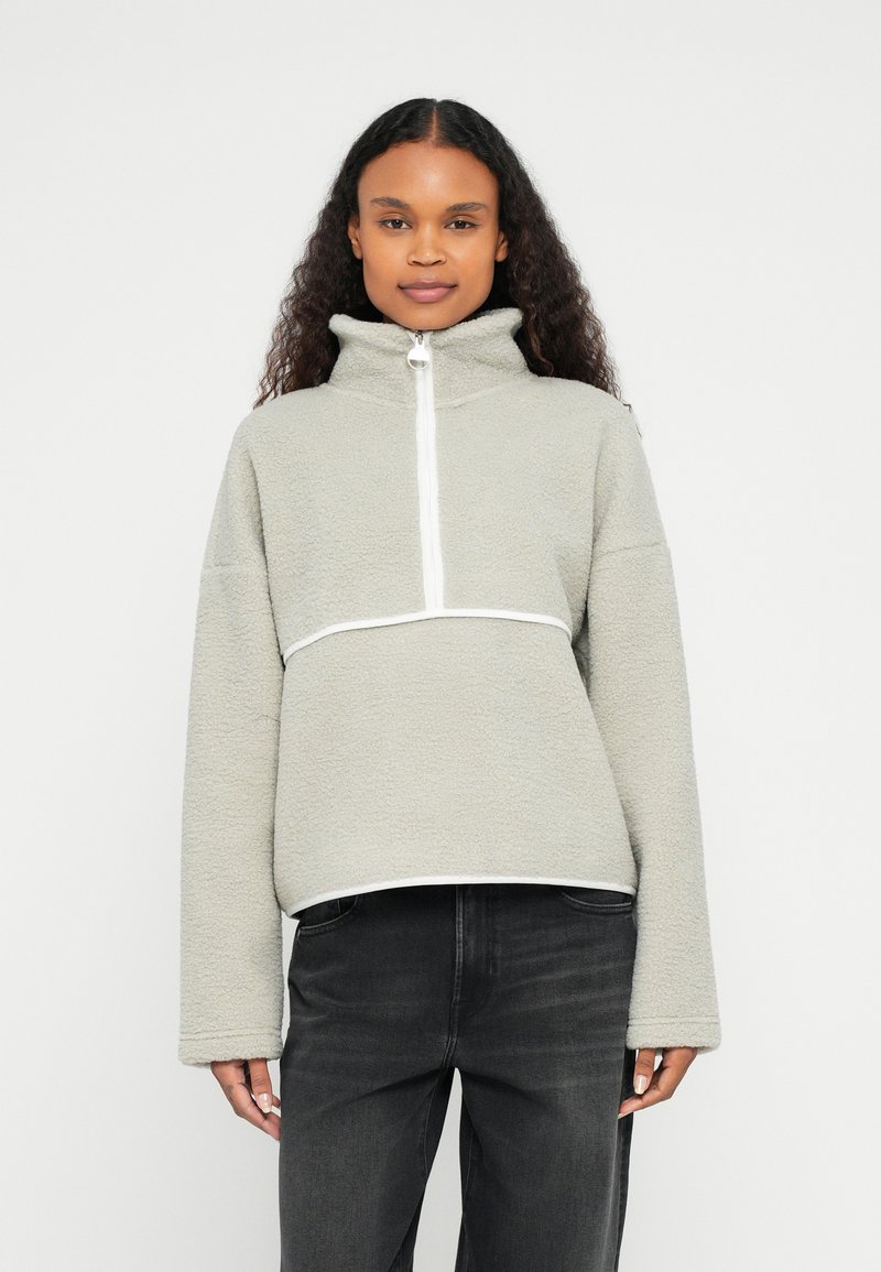 Jasnozielona bluza polarowa z wysokim kołnierzem, suwakiem do ćwiartki i białym akcentem pod klatką piersiową oraz przy dolnym brzegu. Miękka, teksturowana tkanina.