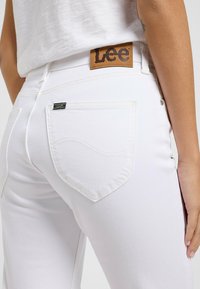 Jeans de denim blanco con un parche de cuero marrón de la marca Lee en la cintura, bolsillos traseros con costura curva y una textura suave.