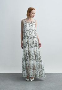 Femme en robe longue blanche sans manches avec broderie florale verte et chaussures blanches à bouts pointus, debout sur un sol gris devant un mur gris clair.