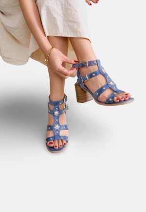 Femme attachant des sandales à talons hauts bleues ornées de clous en forme de soleil et de talons épais en bois, portant une jupe beige clair et du vernis à ongles rouge.