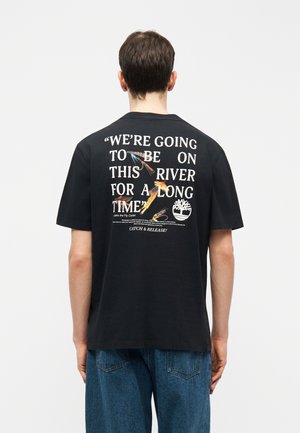 Moški, ki nosi črno majico s belim napisom in barvitim motivom ribolovnih muh, stoji obrnjeno stran proti enostavni ozadju.