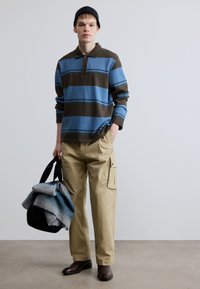 Sinine ja pruun triibuline polo särk, beežid cargo-püksid, mustad kingad ja must kott koos halli ja sinise pleediga.