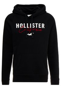 Svart huvtröja i mjukt material med en känguruficka, dragsnören och broderad "HOLLISTER California"-logotyp i vitt och rött.