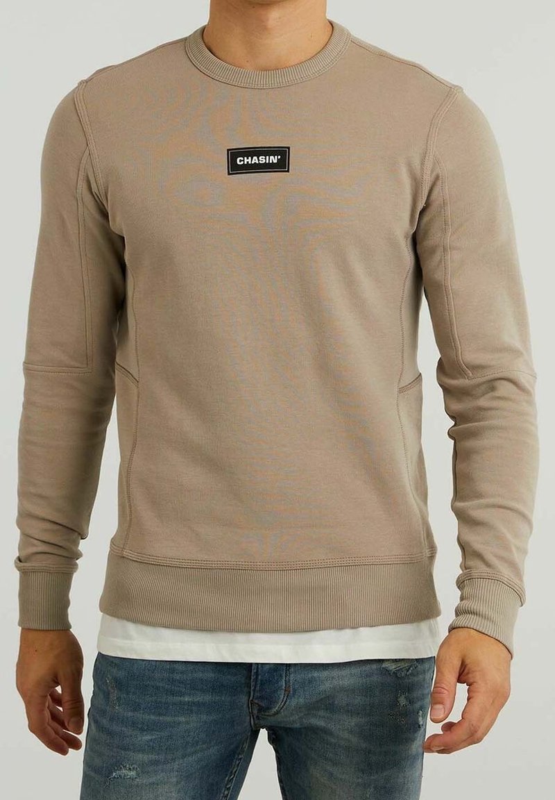 Homme portant un sweat-shirt beige à manches longues avec le logo "CHASIN'" sur la poitrine et un jean bleu, debout devant un fond uni.
