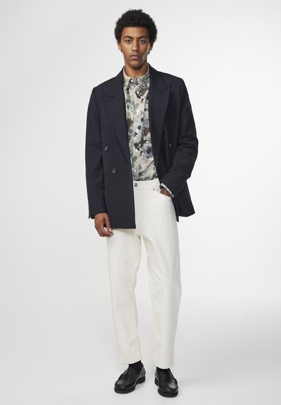 Blazer croisé navy sur une chemise à motifs, associé à un pantalon blanc et des chaussures noires. Présente un design ajusté et des textures lisses.