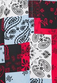 Tissu à motif bandana multicolore comportant des carrés rouges, noirs, blancs et bleu clair avec des motifs paisley et floraux. Texture lisse.