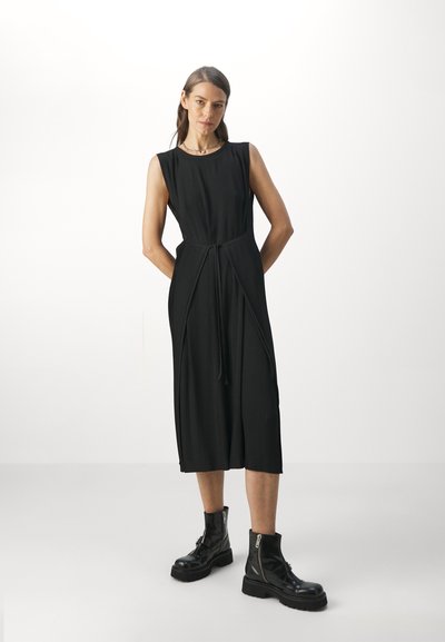 Robe noire sans manches avec une texture plissée, taille cintrée et fentes sur les côtés, associée à des bottines noires remontant jusqu'à la cheville avec des zip et des semelles épaisses.