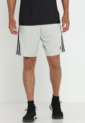 Mann trägt weiße Adidas-Shorts mit schwarzen Streifen und Logo, schwarze Sportschuhe und schwarzes Shirt, steht auf weißem Boden und Hintergrund.