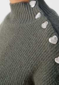 Gros plan d'un pull en maille côtelée grise avec un col montant, doté de cinq boutons argentés en forme de cœur ornés de strass le long de l'épaule.