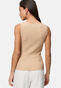 Débardeur beige à côtes avec des bords festonnés aux épaules, design ajusté et tissu texturé, porté avec un pantalon de couleur claire.