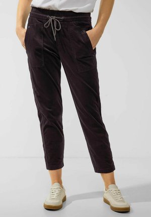 Dunkelviolette Cordhosen mit elastischem Bund und Kordelzug, seitlichen Taschen und schmal zulaufendem Schnitt. Getragen mit beigen Sneakers.