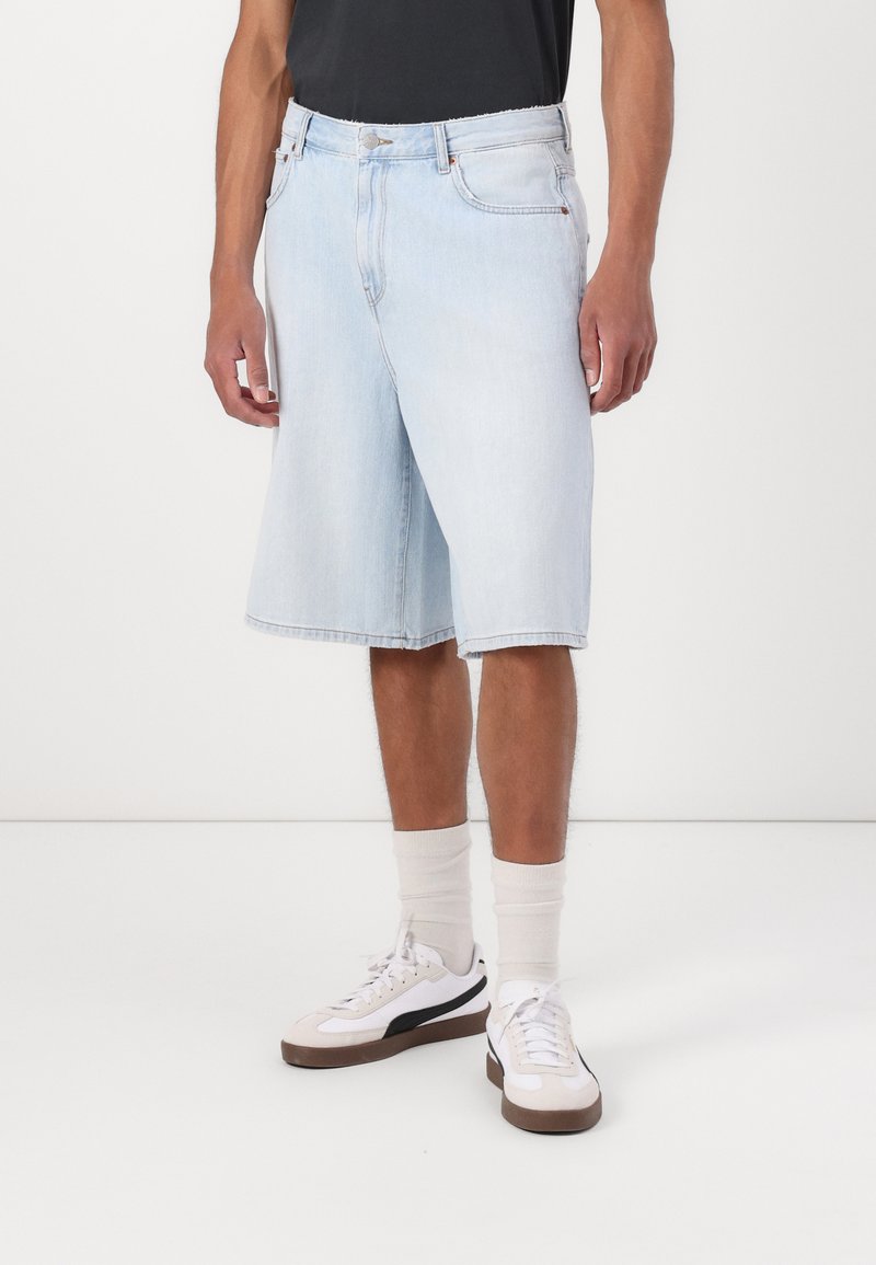 Shorts en denim bleu clair avec une coupe ample, arrivant au genou, dotés d'une fermeture à bouton et de poches. Portés avec des baskets blanches et des chaussettes crème.