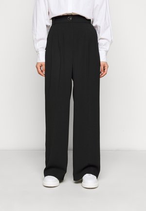 Person wearing high-waist black wide-leg trousers with front pleats, white button-up shirt, and white sneakers standing against a plain white background.