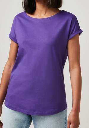 Vrouw draagt een effen paarse korte mouwen shirt met opgerolde manchetten, gecombineerd met lichtblauwe jeans, staande tegen een effen achtergrond.