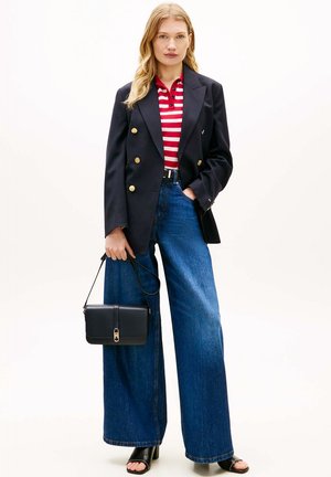 Vrouw draagt een navy blazer met gouden knopen, een rood-wit gestreept overhemd, wijde blauwe jeans, zwarte sandalen met hak, en houdt een zwarte handtas vast.