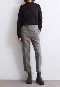 Pull noir en maille avec col montant et épaules tombantes, associé à un pantalon gris à carreaux et des bottines noires à semelle épaisse.
