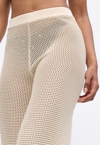 Beige verkko leggingsit korkealla ribbaus vyötäröllä, joissa on avoin neulekuvio ja sileä pinta. Lähikuva jalasta ja kädestä.