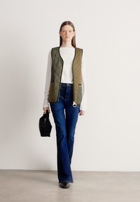 Pihe-puha zöld mellény fehér ing felett, sötétkék bőszárú farmerral párosítva. A modell egy fekete táskát tart, világosszürke padlón áll.