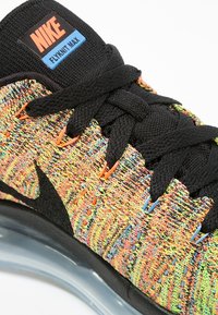 Gros plan de la basket Nike Flyknit Max montrant un tissu tricoté multicolore, des lacets noirs, le swoosh Nike noir et une partie de la semelle transparente.