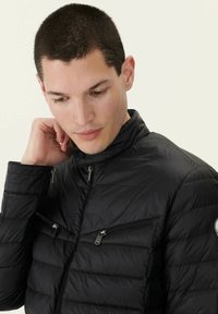 Giacca nera isolante con colletto alto, tasche con zip e texture a righe orizzontali. Materiale leggero con una finitura liscia.