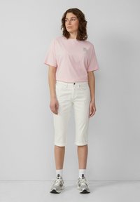 T-shirt rose clair à manches courtes avec un logo. Jean blanc, coupé, avec des ourlets retroussés et des baskets argentées, accompagné de chaussettes blanches.