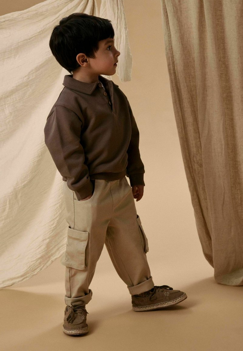 Brown long-sleeve polo with three buttons, paired with beige cargo pants featuring side pockets, and dark shoes with a light sole.