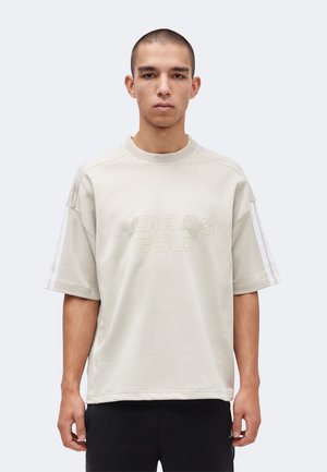 Junger Mann mit kurzen Haaren trägt ein beiges Adidas Golf T-Shirt mit erhabenem Logo und schwarze Hose, steht vor einem schlichten weißen Hintergrund.