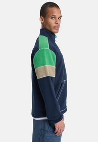 Φλις πουλόβερ σε ναυτικό μπλε με πράσινες και μπεζ λεπτομέρειες, μισό φερμουάρ στον λαιμό και υφή υφάσματος, με άνετη γραμμή και πλαϊνή τσέπη.