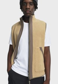 Beige Fleece-Weste mit vollem Reißverschluss, braunen Akzenten und Seitentasche. Über einem weißen T-Shirt getragen, zeigt sie eine weiche Textur und ein lässiges Design.