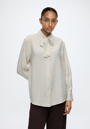 Donna con capelli scuri in camicetta beige a maniche lunghe con dettaglio a fiocco al collo e pantaloni neri, in piedi su uno sfondo bianco uniforme.