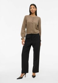 Une femme se tient en portant un cardigan marron avec boutons, un pantalon noir taille haute large, et des escarpins noirs à bout pointu sur un fond blanc.