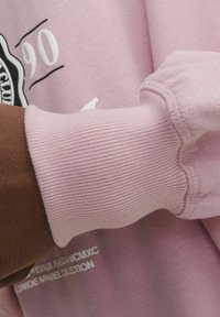 Sudadera rosa claro con puños acanalados. Presenta un estampado gráfico y una textura suave. Primer plano del detalle de la manga.