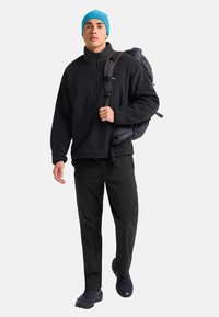 Jonge man in zwarte fleece, zwarte broek, blauwe beanie die een donkergrijze rugzak over één schouder draagt, loopt naar voren op een witte achtergrond.