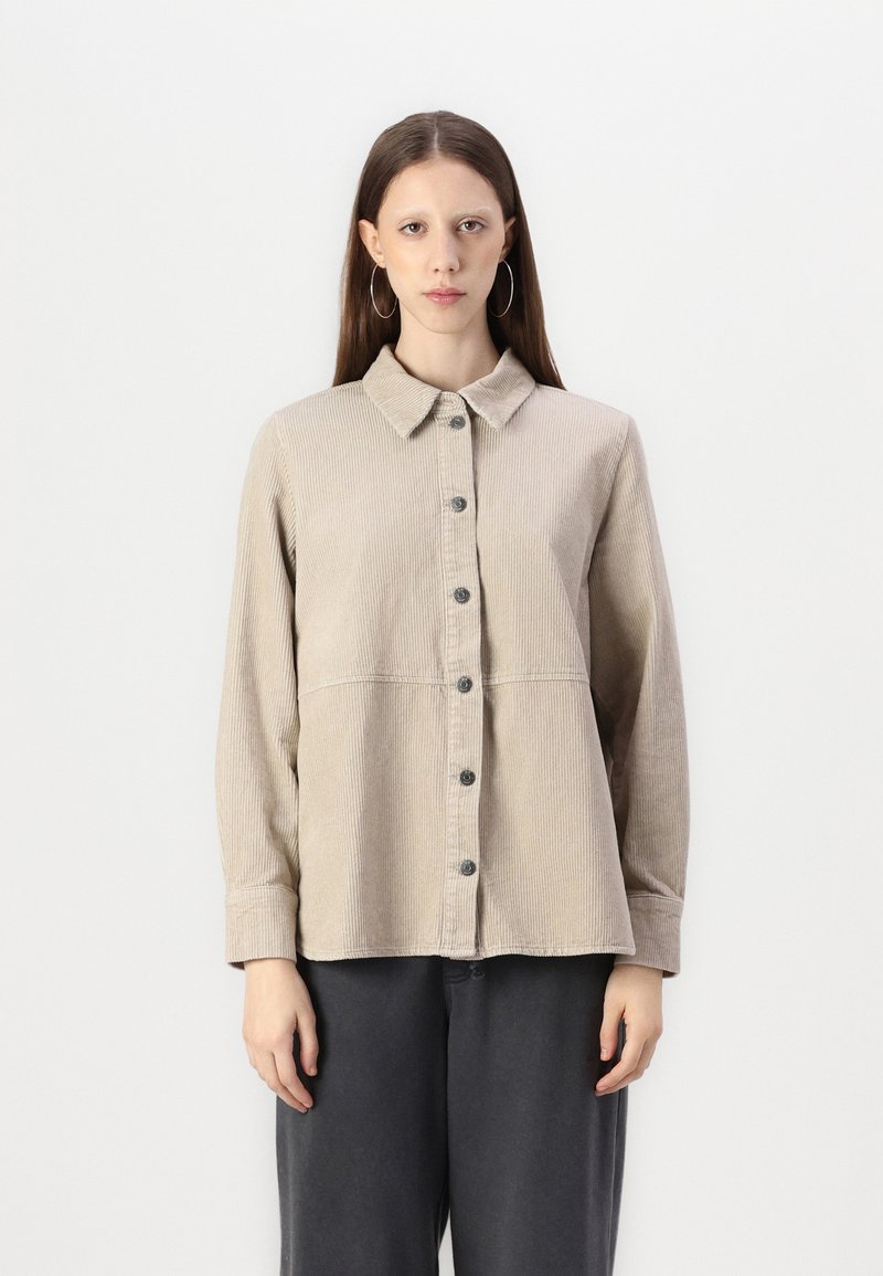 Beige kordfløyelskjorte med krage, knapper foran, lange ermer og en horisontal søm over torsoen. Teksturert stoff.