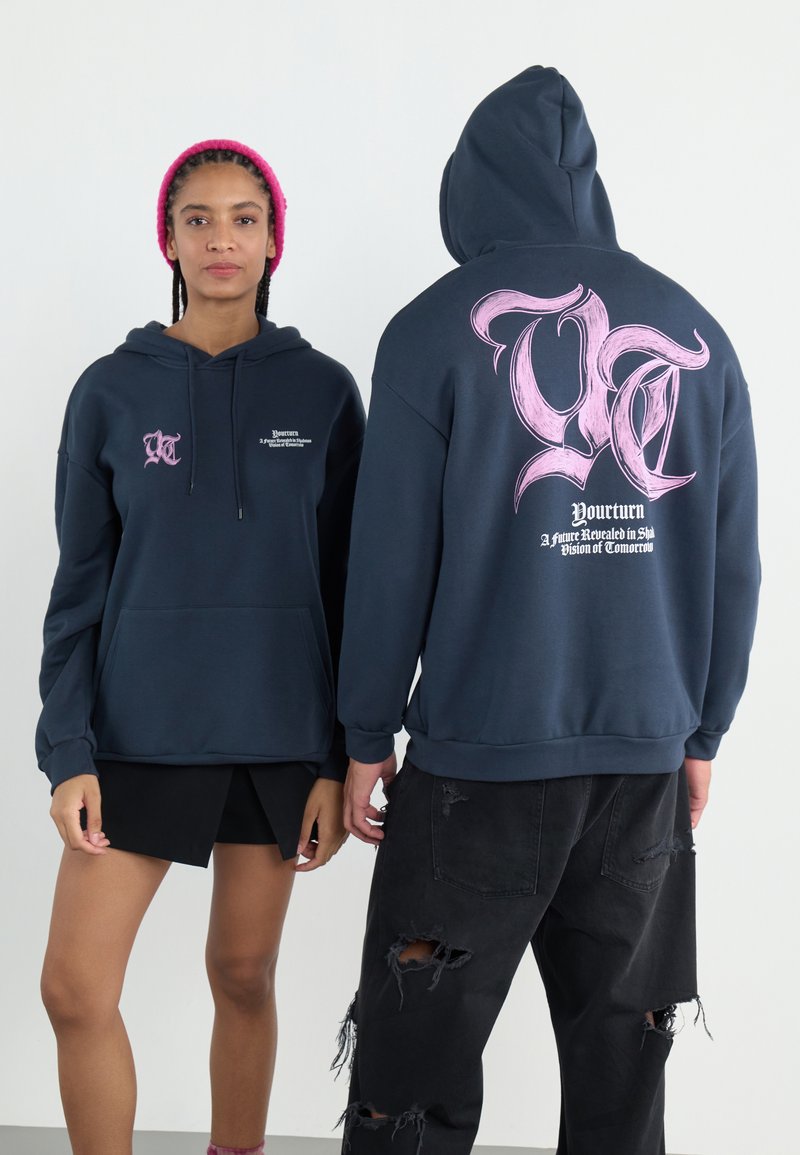 Marineblå hettegenser med frontlomme, med en rosa grafisk logo på baksiden og trykt tekst. Stoffet ser mykt og teksturert ut.