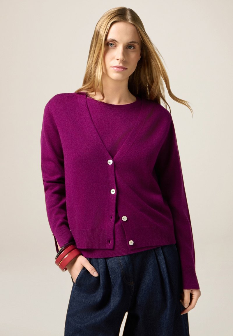 Cardigan viola con scollo a V, chiusura con bottoni e orlo a coste. Abbinato a pantaloni navy a gamba larga e braccialetti rossi al polso.