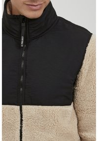 Black and tan fleece jacket with a high collar, featuring a zip closure and quilted texture on upper section. Soft, shaggy lower panel.