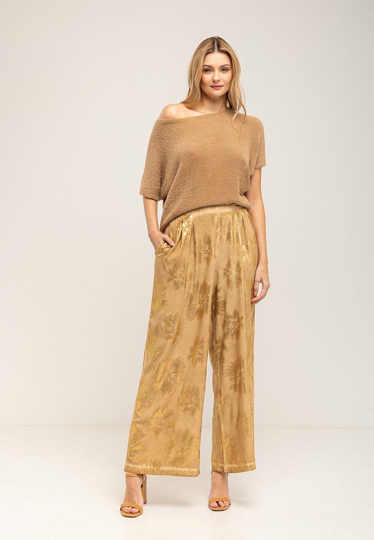 Kobieta w luźnym brązowym swetrze z opuszczonymi ramionami, szerokich złotych spodniach w kwiaty oraz dopasowanych sandałach na obcasie, stoi z jedną ręką w kieszeni.